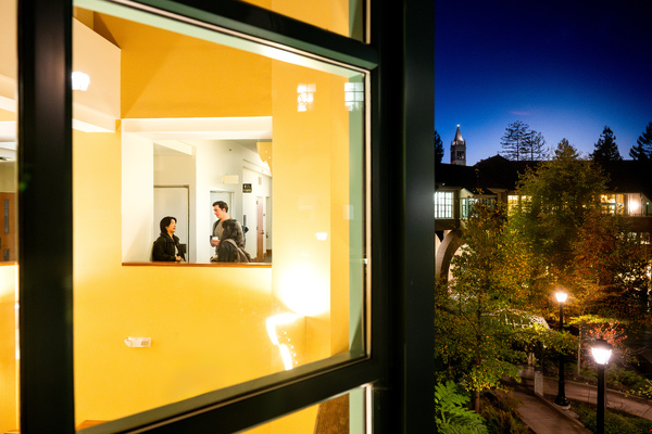 View through a window into a warmly lit hallway where a few people are talking near a classroom doorway at night. Outside, the UC Berkeley campus is visible under a deep blue sky, with the Campanile tower illuminated in the distance and lamplight reflecting off trees and pathways below.